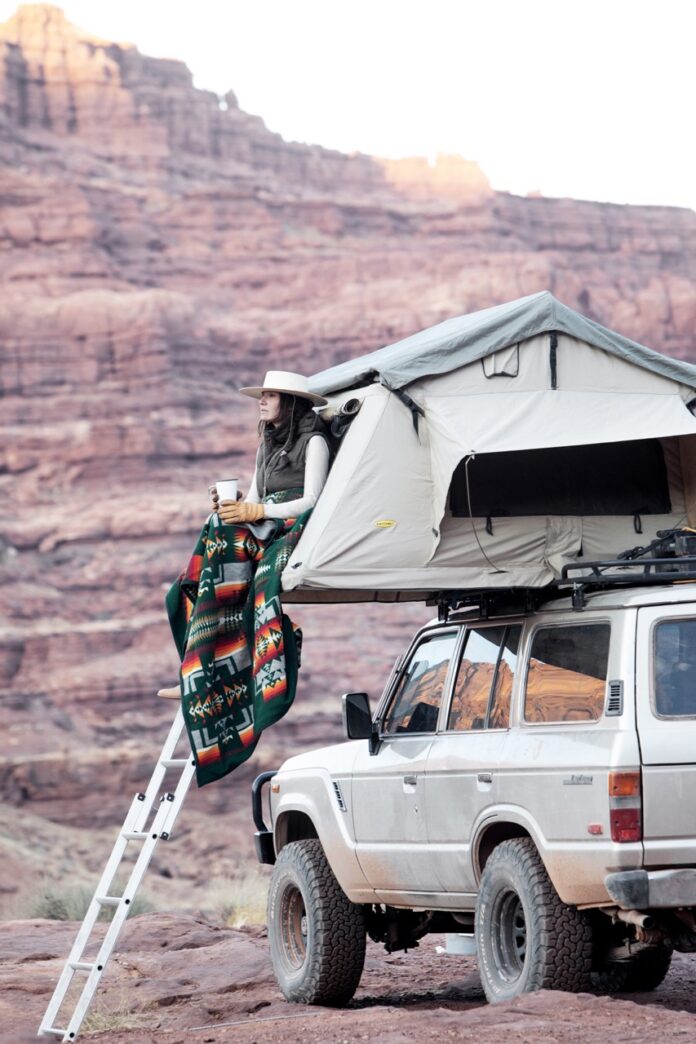 A woman sitting on the upper tent on top of a truck while glamping.