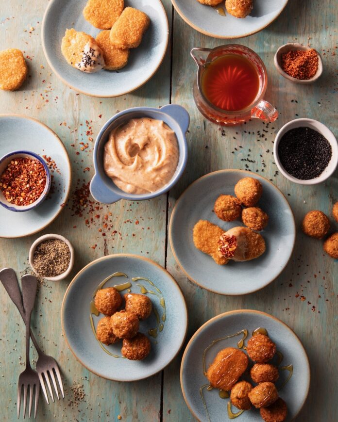 Various small plates of appetizers like chicken nuggets and falaffel surround a small bowl of Hot Honey Aioli with Harissa.