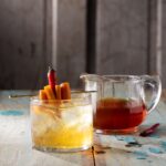 A short glass holds a Hot Honey Old Fashioned with an orange slice and chili pepper garnish as a small pitcher of hot honey sits off to the right.