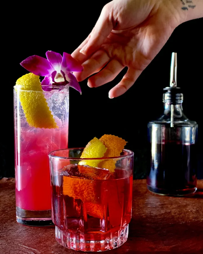 How to Make Hibiscus Cocktails- Bright, Floral Drinks from a Pittsburgh Distiller Two Hibiscus cocktails, one in a rocks glass, one in a high ball glass. The later is very pink in color and a white had is putting a pink flower on top.