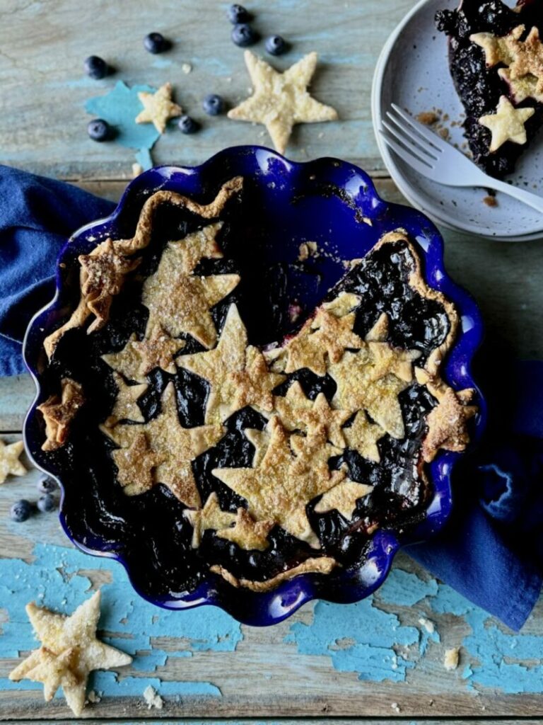 A cherry blueberry pie with stars as a top crust, in a cobalt blue, scalloped edge pie dish sitting on a blue cotton towel on a rustic wooden surface with chipped light blue paint