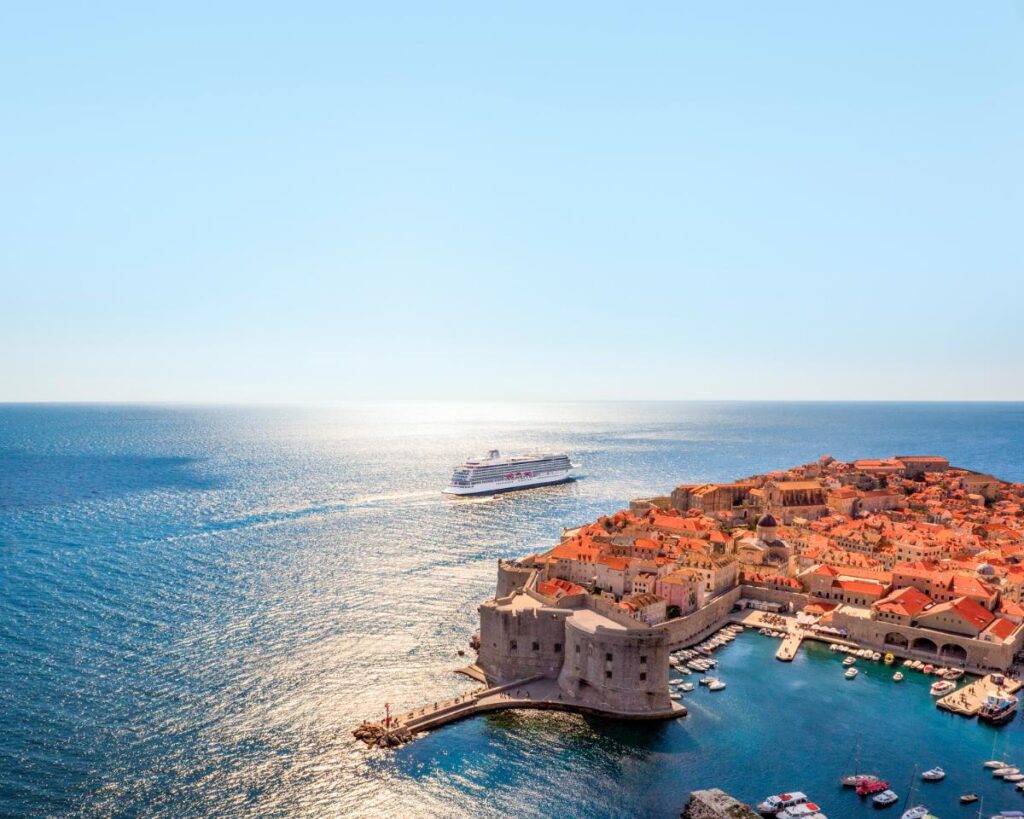 An aerial view captures a large white cruise ship sailing on a blue sea next to a coastal town with red-roofed buildings.