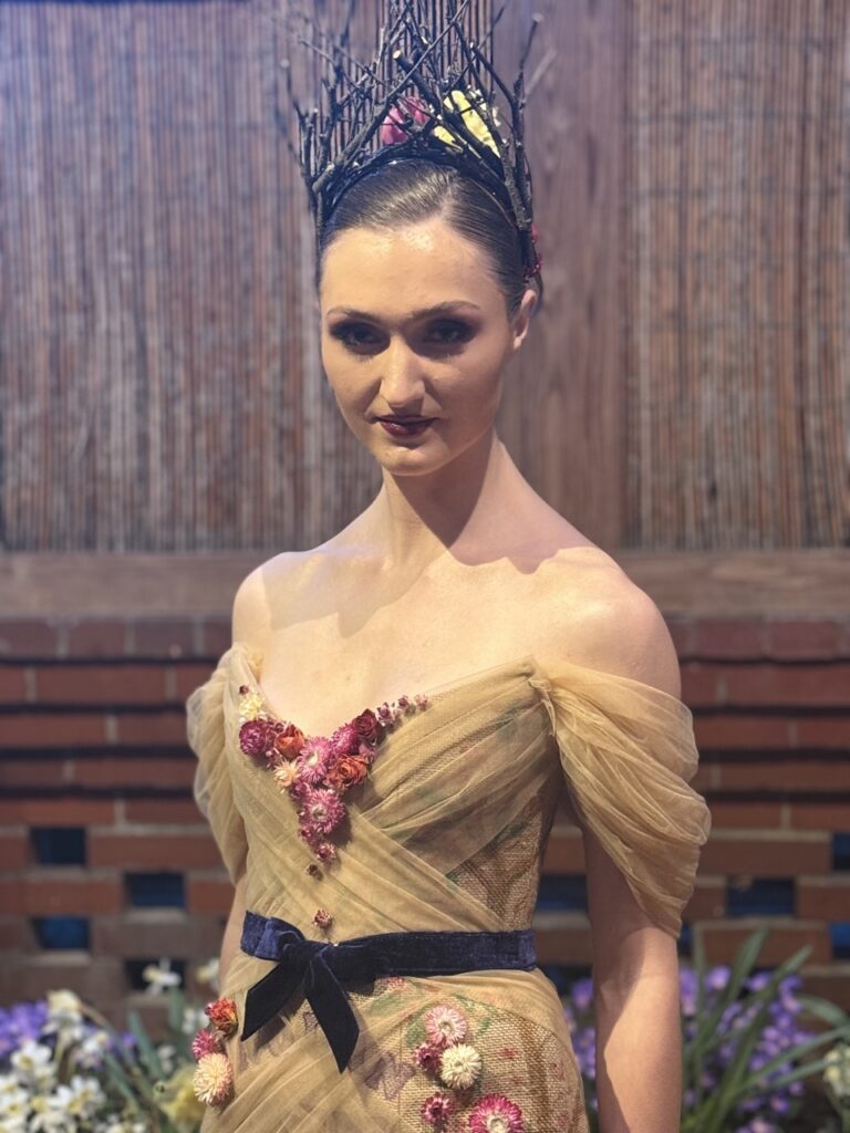 A woman in a brown dress stands against a wall.