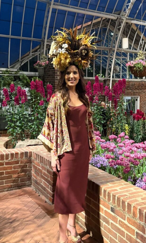 A woman in a red dress and cardigan stands in front of florals.