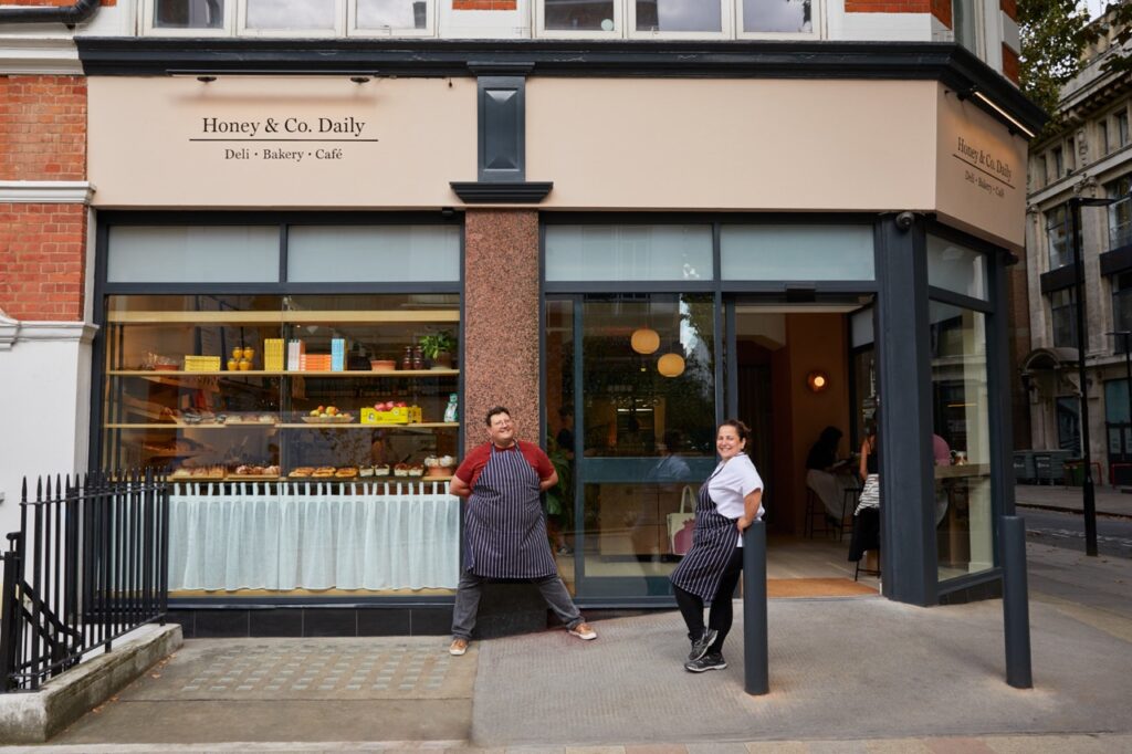 A man and woman in aprons stand outside of Honey and Co restaurant in London.