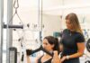 Luxury Fitness in Pittsburgh to Become Your Best Self A woman supports another woman as she works out at a pull down machine in a luxury fitness gym.