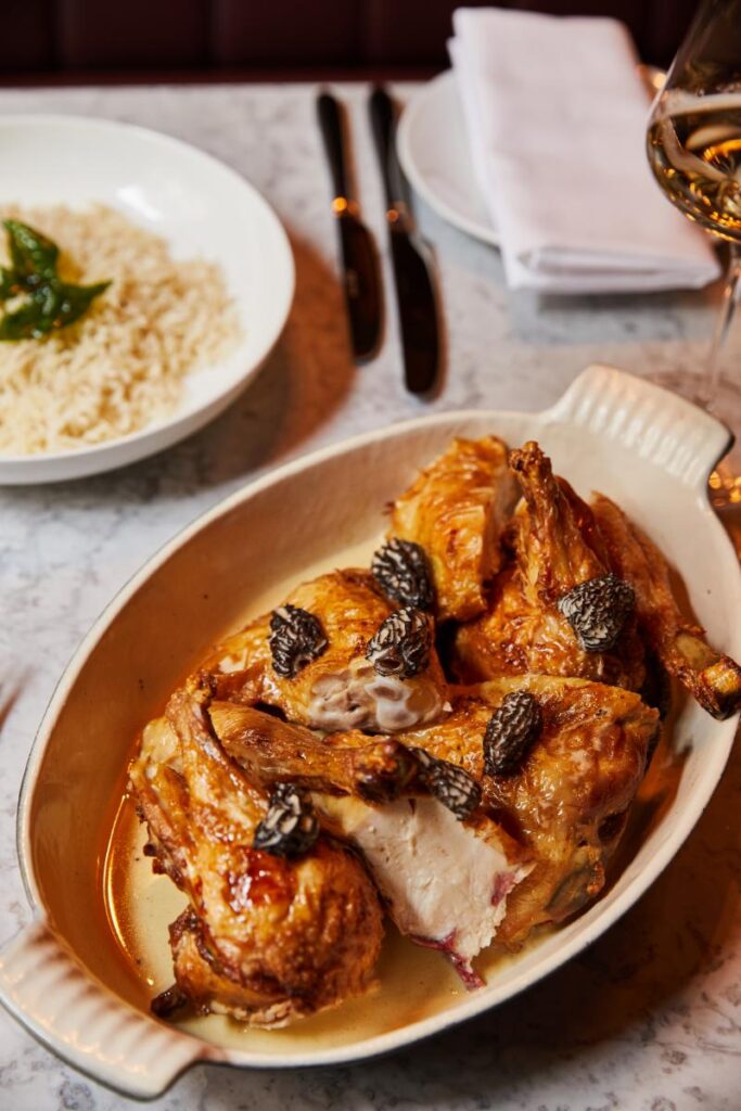 A close-up of a perfectly cooked roast chicken, sliced and served in a white baking dish, garnished with morels.