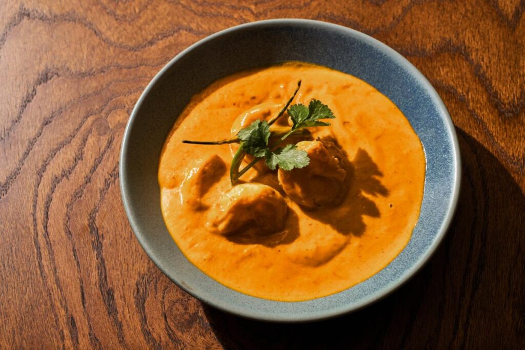 A vibrant orange prawn malaikari curry with three large prawns, garnished with cilantro, served in a blue bowl on a wooden table.