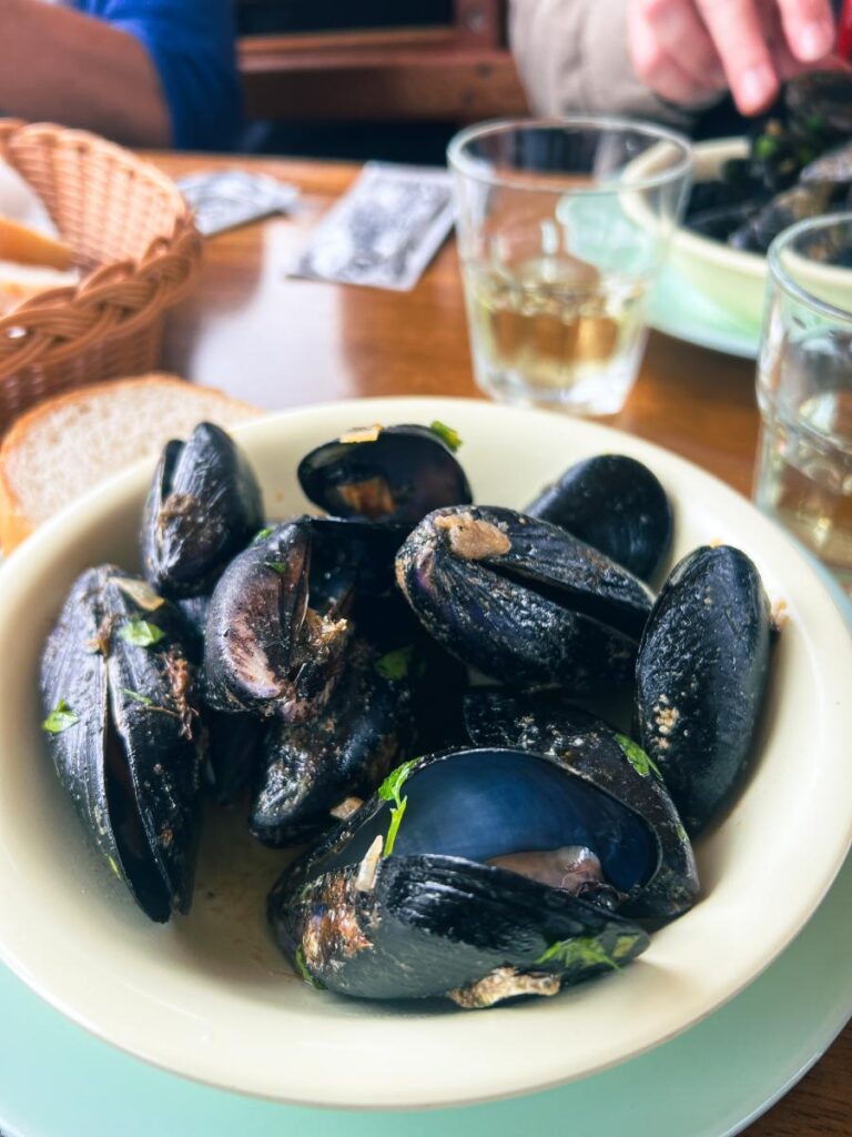 A plate of mussels in their shells, garnished with green herbs, is centered on a light-colored table with a glass of white wine and a bread basket in the background.