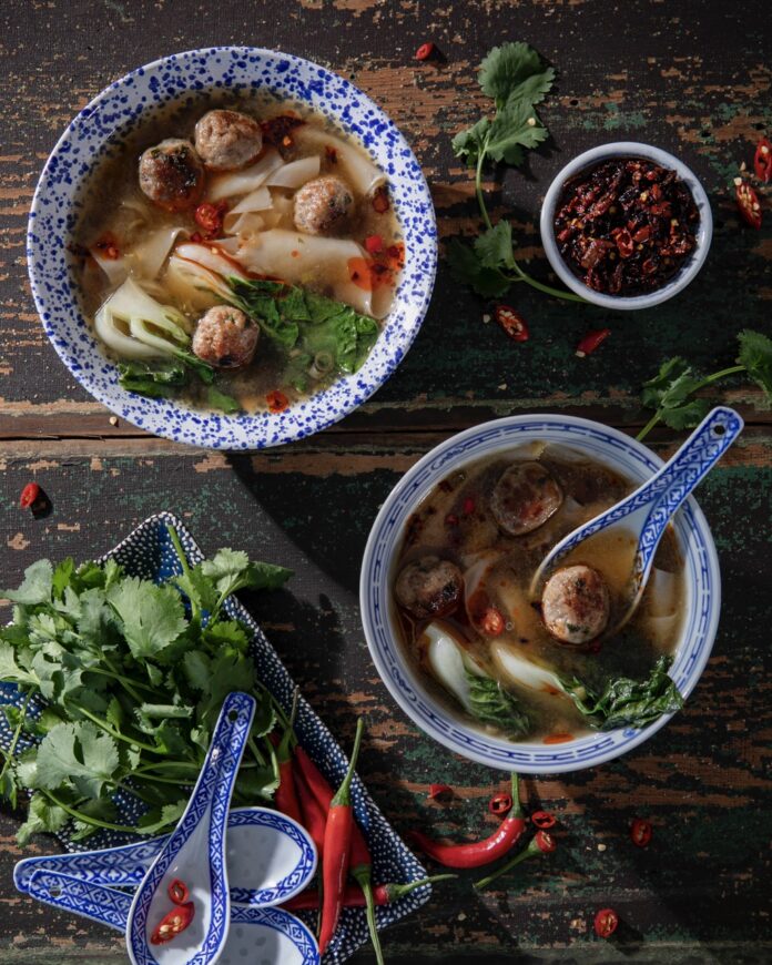 A large blue and white bowl of quick and easy wonton soup sits diagonal to a smaller bowl that has a wonton soup spoon inside it. Both bowls are surrounded by greens and chili peppers.