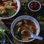 A bowl of quick and easy pork wonton soup in a blue china bowl with a blue pattern dumpling spoon holding a pork ball.