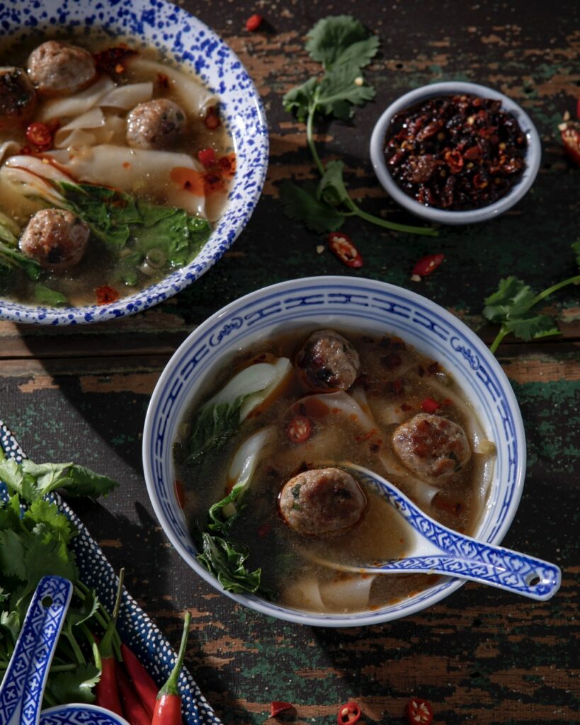 A bowl of quick and easy pork wonton soup in a blue china bowl with a blue pattern dumpling spoon holding a pork ball.