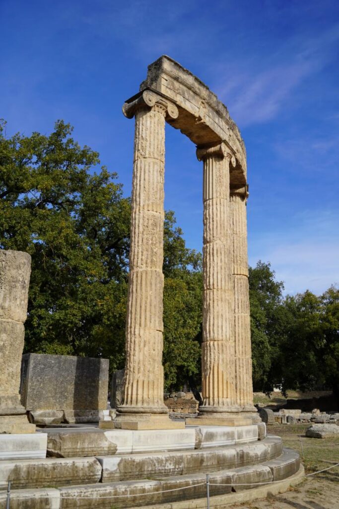 Two large, fluted stone columns stand upright against a bright blue sky, with green trees in the background.