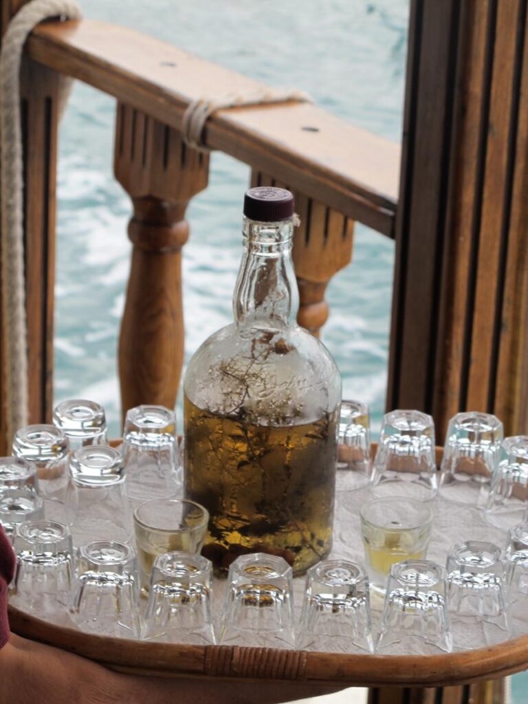 A large, clear bottle filled with amber liquid and a few submerged herbs stands on a white tray surrounded by many small, empty glasses on a boat railing.