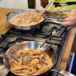 On a stovetop, a hand with tongs reaches into a steel pan filled with head-on shrimp to make 10 Minute Aegean-Style Shrimp with Caper Almond Pesto.