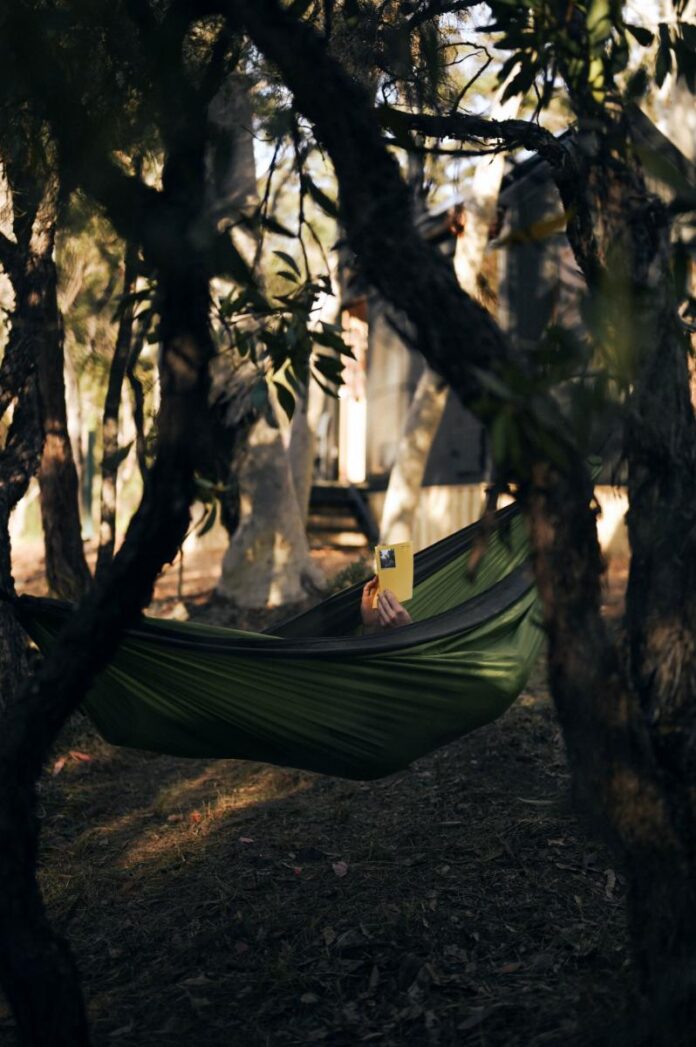 A dark green hammock strung between trees in a forest. The person is holding a bright yellow book.
