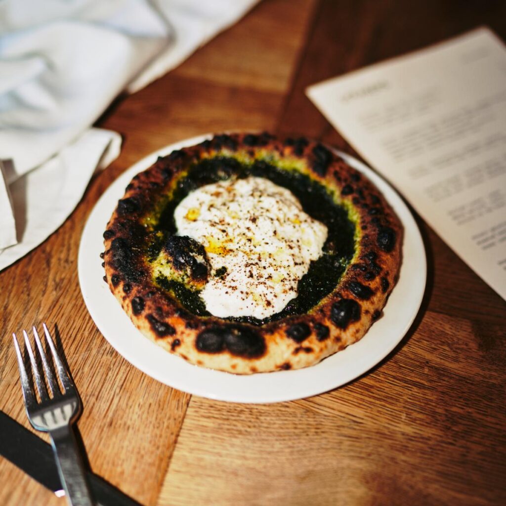 A flatbread with a slightly charred crust, topped with a large dollop of white burrata cheese and green herbs, served on a white plate with a fork and knife.