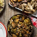An aerial shot of a wooden surface with plates of food, the bottom right, in a cast iron pan, sits Pittsburgh BBQ Recipes from Gaucho Parrilla Argentina: Chipotle Brussels Sprouts.