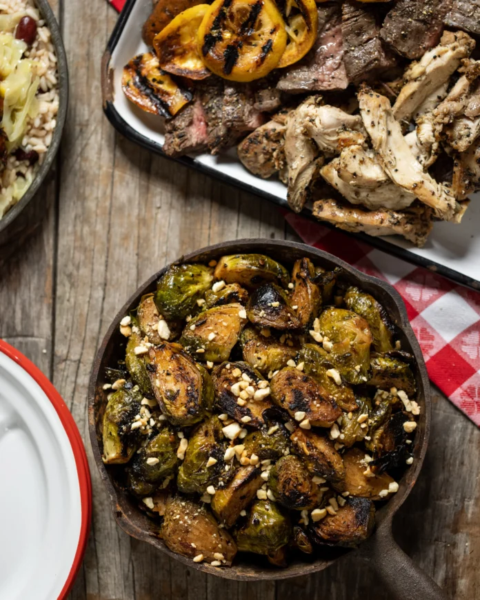 An aerial shot of a wooden surface with plates of food, the bottom right, in a cast iron pan, sits Pittsburgh BBQ Recipes from Gaucho Parrilla Argentina: Chipotle Brussels Sprouts.