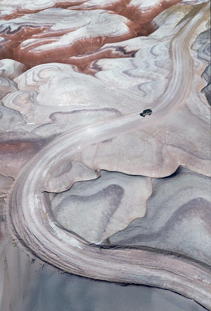An aerial shot of a dark-colored SUV driving on a winding dirt road through a vast, undulating landscape with layers of red, white, and grey rock formations.