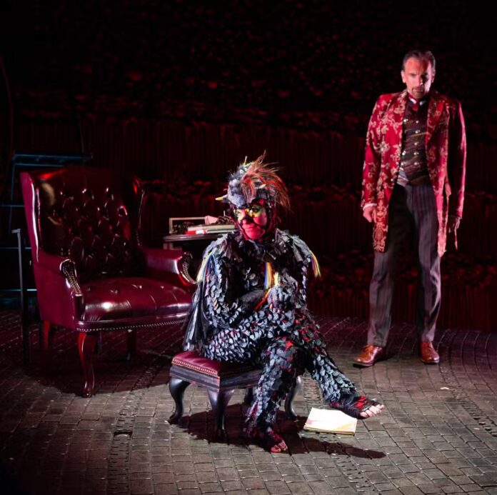 Screwtape Letters A costumed actor in The Screwtape Letters in Pittsburgh sits on a small chair on stage as the devil watches from the back.