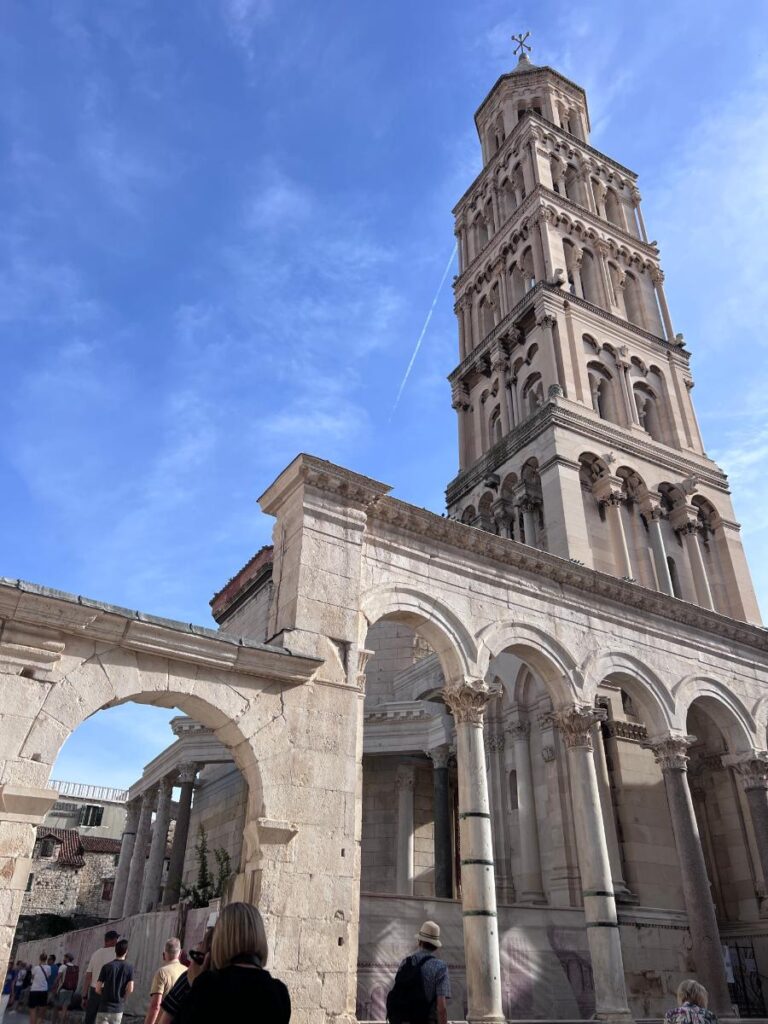 A towering, light-colored stone bell tower with multiple arched levels stands against a bright blue sky with wispy clouds.
