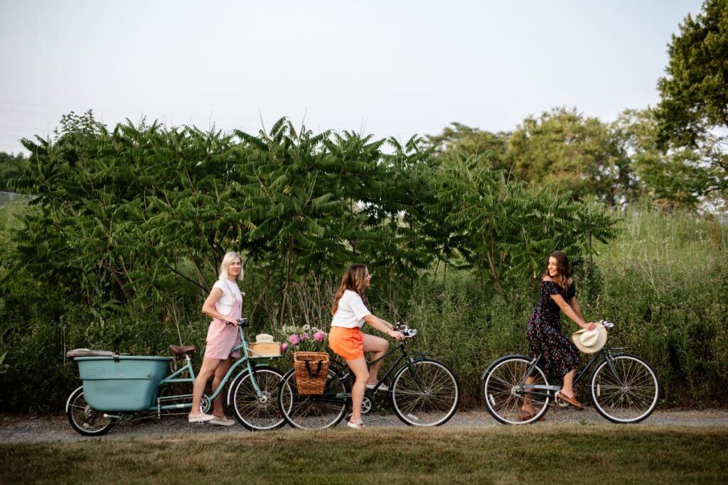 Three sisters ride bicycles on a dirt path next to green foliage.