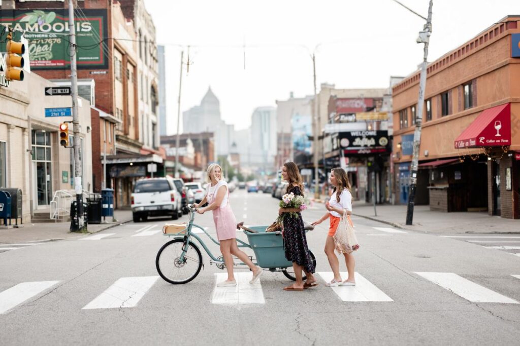 Three sisters walk across a crosswalk in a city, with one pushing a light blue cargo bike.