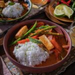 Two bowls of 20 minute coconut red curry with rice and vegetables sit on a table beside fresh limes and beer.