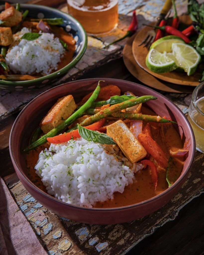 Two bowls of 20 minute coconut red curry with rice and vegetables sit on a table beside fresh limes and beer.