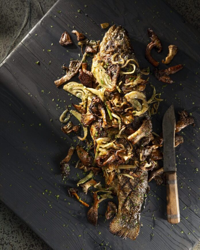 On a black wooden surface is a Rainbow Trout with Fennel and Mushrooms sitting next to a knife.