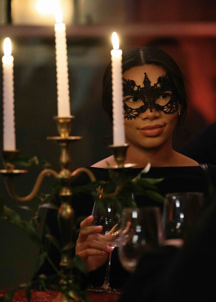 A woman with a lace eye mask standing behind a candelabra with 3 lit white candles in a room washed in red lighting.
