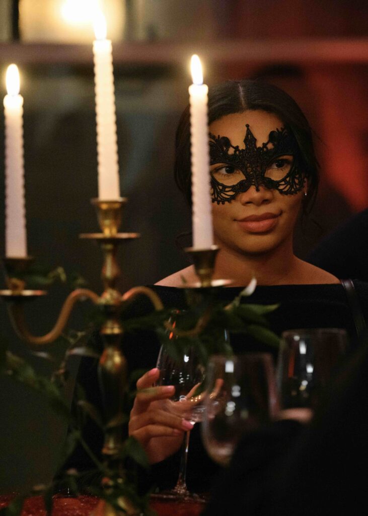 A woman with a lace eye mask standing behind a candelabra with 3 lit white candles in a room washed in red lighting.