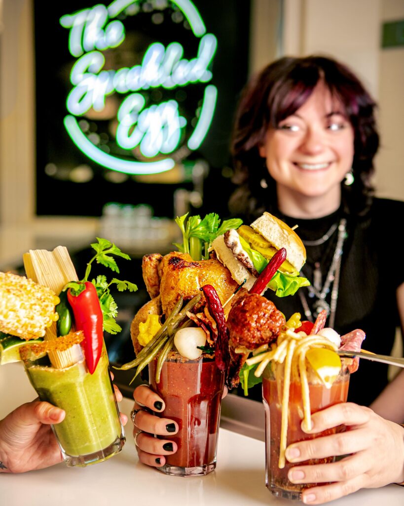 Three loaded bloody Marys in people's hands in from of a woman behind a bar at The Speckled Egg in downtown Pittsburgh.