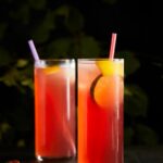 Two tall highball glasses with a reddish-pink cocktail and a colorful straw, garnished with fruit slices.