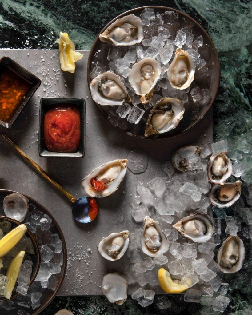 A top-down view of raw oysters on the half shell served on ice, with two different dipping sauces, lemon wedges, and a serving spoon.