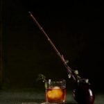 A Coffee Old Fashioned cocktail in a rocks glass with a large ice cube, garnished with a leafy branch, next to a caramel apple on a stick.