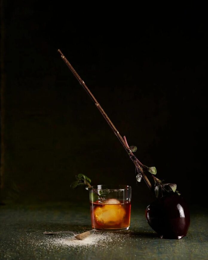 Coffee Old Fashioned A Coffee Old Fashioned cocktail in a rocks glass with a large ice cube, garnished with a leafy branch, next to a caramel apple on a stick.
