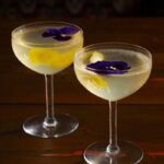 Two cocktails in coupe glasses on a wooden table, each garnished with a lemon peel and a purple edible flower.