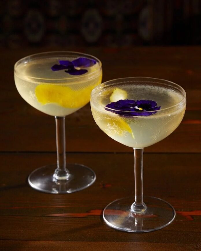 Two cocktails in coupe glasses on a wooden table, each garnished with a lemon peel and a purple edible flower.