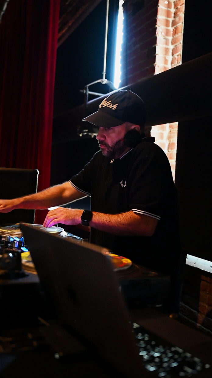 A DJ spinning records wearing a ball cap in a darkly lit room.