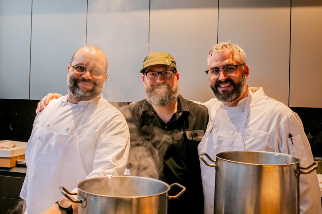 Three chefs stand behind pots of water.