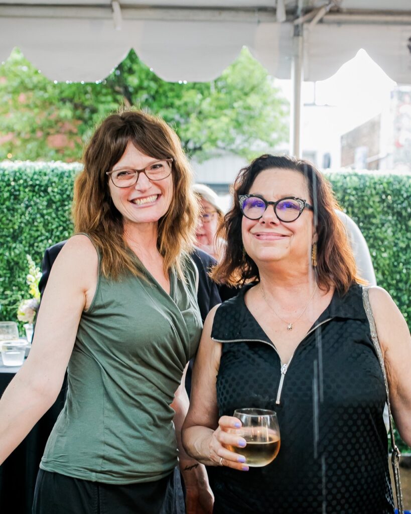 A woman in a green shirt and a woman in a black shirt both with glasses stand together.