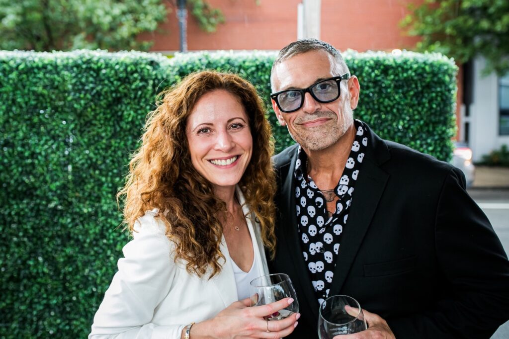 A man in a black suit jacket and skull shirt stands with a woman in a white jacket.