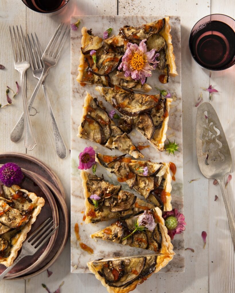 An eggplant and smoked mozzarella tart cut into pieces on a white table beside silverware and plates.
