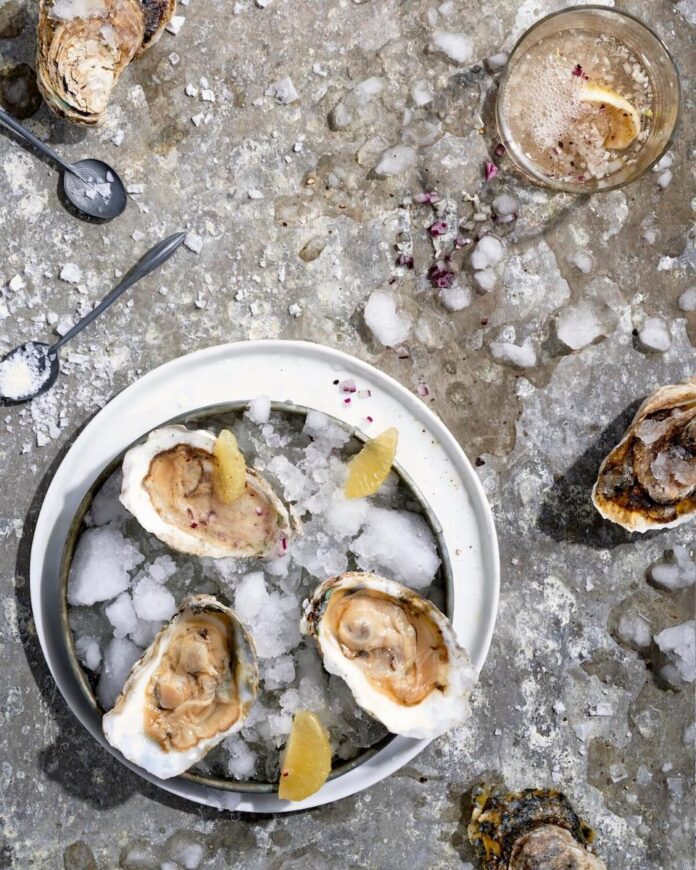 Several oysters on the half shell served on ice, with lemon wedges and a drink nearby.