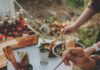 Recipes for a Farm to Table, Vegan Tailgate A hand holds a pasta dish from a buffet table, featuring pulled pork, green beans, and other fresh vegetables.