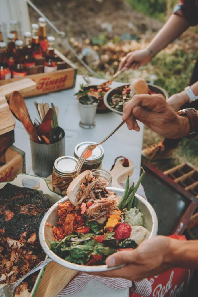A hand holds a pasta dish from a buffet table, featuring pulled pork, green beans, and other fresh vegetables.