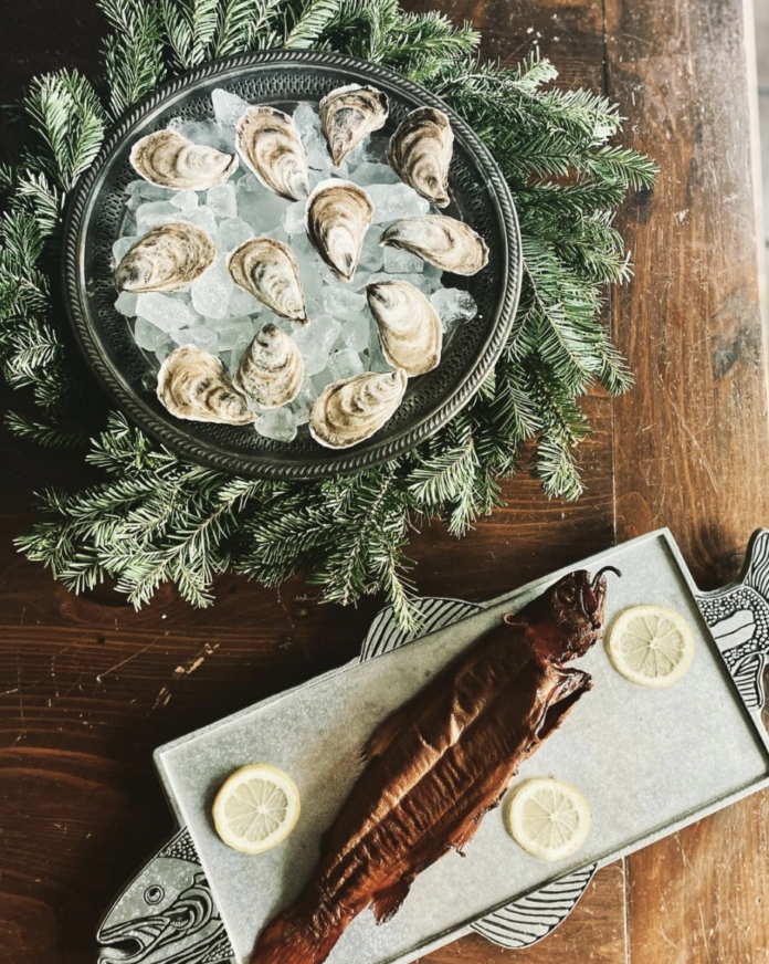 A platter of oysters on the half shell served on ice, surrounded by evergreen branches, next to a smoked fish with lemon slices.