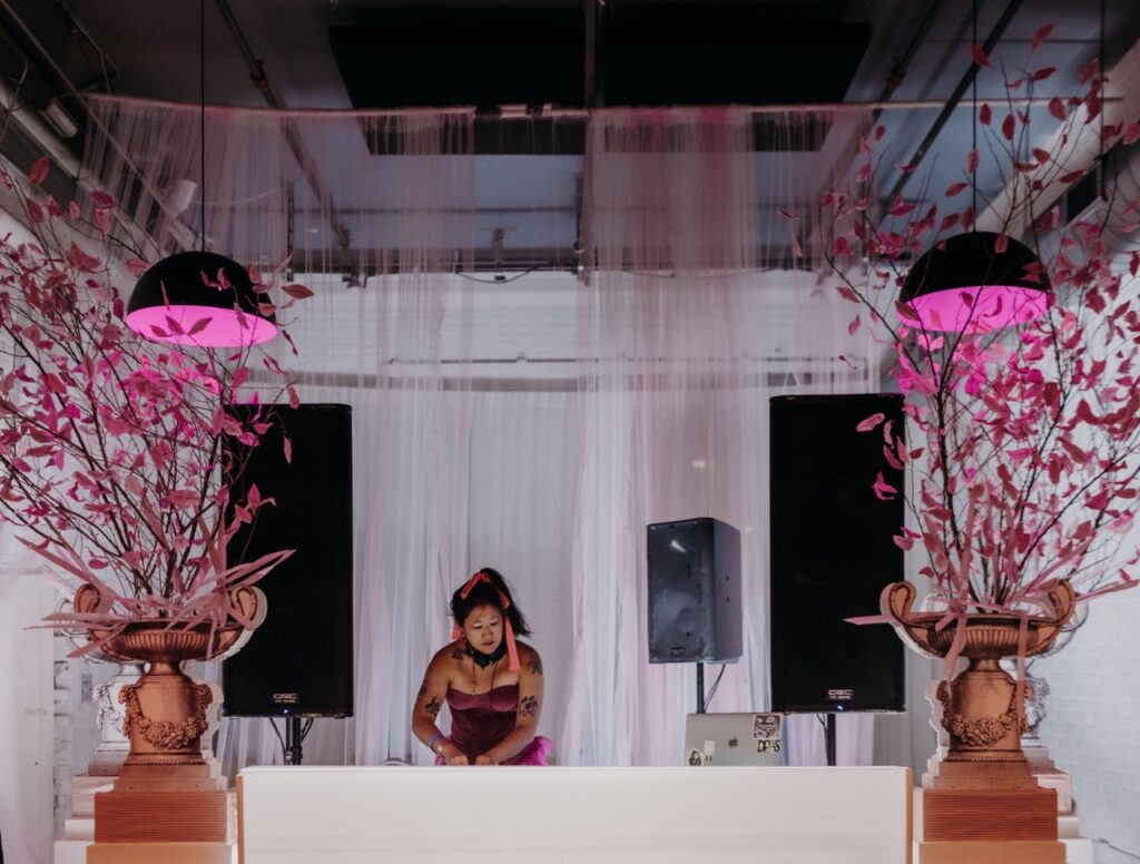 A woman in a pink dress DJs between two pink lights and trees.